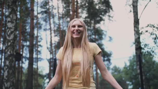 Glad kvinna hoppar på studsmatta. Vacker ung blond barfota kvinna i ljus sommar skjorta och jeans tittar på kameran och ler hoppa på stor studsmatta med höga träd runt. — Stockvideo