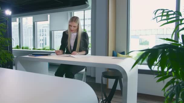 Femme élégante travaillant dans un bureau élégant. Femme blonde moderne en costume tendance assise à table dans un bureau contemporain léger et écrivant sur des papiers .