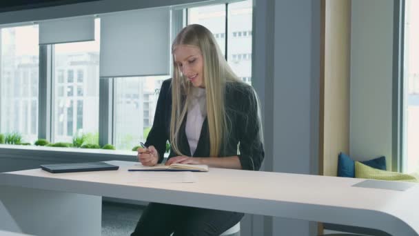 Donna elegante che lavora in ufficio elegante. Moderna donna bionda in abito alla moda seduta a tavola in leggero ufficio contemporaneo e scrittura su carta . — Video Stock