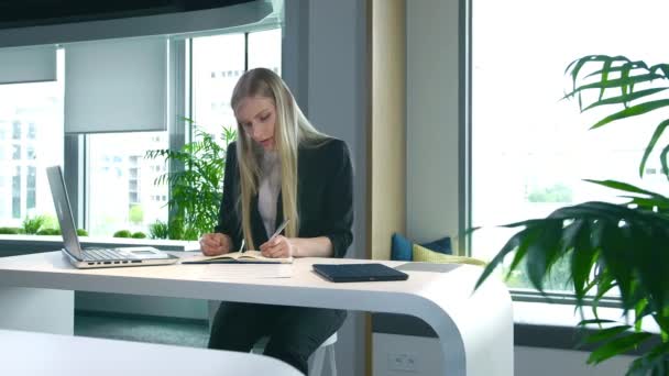 Femme élégante travaillant dans un bureau élégant. Femme blonde moderne en costume tendance assise à table dans un bureau contemporain léger et écrivant sur des papiers . — Video