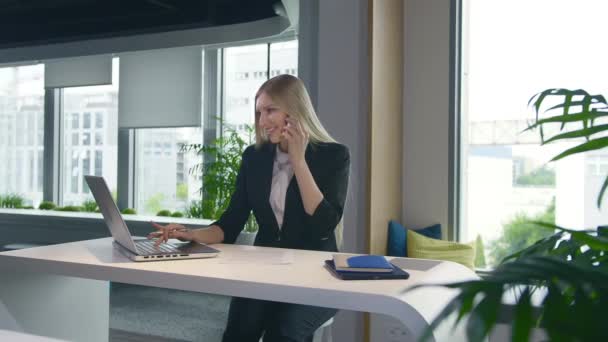 Mujer de negocios de moda viendo el ordenador portátil y hablando por teléfono. Elegante joven rubia que trabaja en la oficina en el ordenador portátil de surf de mesa y tener llamada telefónica mientras sonríe . — Vídeos de Stock