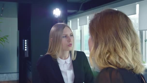 Coworking women speaking in office. Two adult formal women in suits having conversation in modern light office with lamp burning behind. — Stock Video