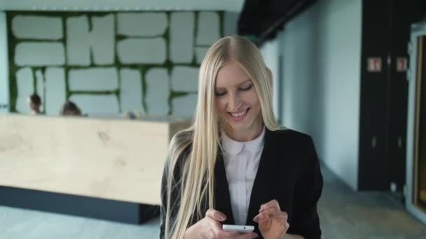 Jovencita alegre con teléfono en la oficina. Mujer bastante rubia con el pelo largo usando un elegante traje negro y sonriendo encantadoramente a la cámara dentro de la oficina moderna . — Vídeo de stock