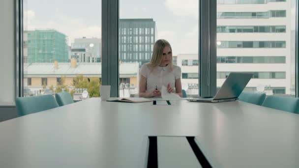 Joven mujer de negocios en la sala de conferencias con papeles. Elegante mujer rubia sentada en una mesa larga en el moderno salón de la oficina con computadora portátil y escritura en papeles mientras trabaja . — Vídeos de Stock