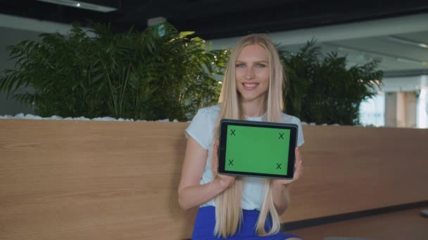 Mulher mostrando tablet no escritório. Jovem mulher loira sorridente mostrando tablet com tela cromada verde olhando para a câmera e sorrindo no escritório . — Vídeo de Stock