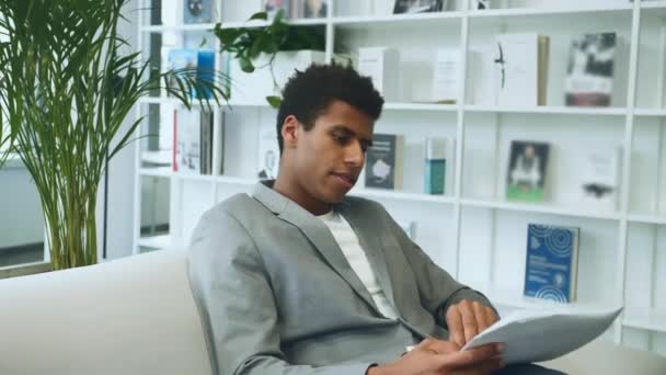 Černý obchodník čte noviny na pohovce. Dospělý Afroameričan v obleku sedí na pohovce v kanceláři a prohlíží si papírové dokumenty. — Stock video