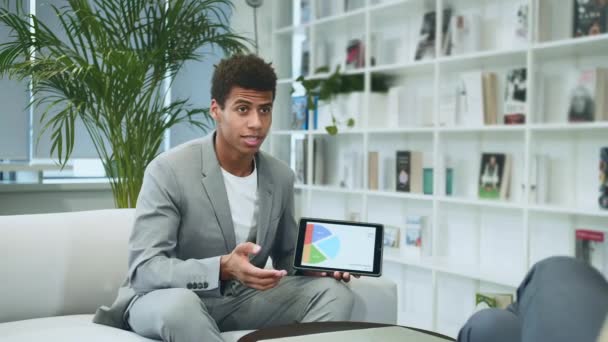Hombre formal negro haciendo presentación con la tableta. Joven hombre afroamericano de traje sentado en la oficina en el sofá con un colega anónimo y haciendo la presentación con la tableta . — Vídeos de Stock