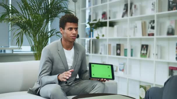 Hombre formal negro haciendo presentación con tableta con pantalla cromakey verde. Joven hombre afroamericano de traje sentado en la oficina en el sofá con un colega anónimo y haciendo la presentación con la tableta . — Vídeo de stock