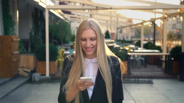 Mujer formal de negocios caminando por la calle. Elegante mujer rubia en traje y caminando por la calle y navegando por el teléfono inteligente con sonrisa contra el fondo urbano . — Vídeos de Stock