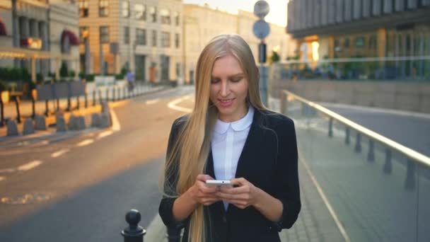 Formellt affärskvinna som går på gatan. Elegant blond kvinna i kostym och promenader på gatan och surfa smartphone med leende mot urban bakgrund. — Stockvideo