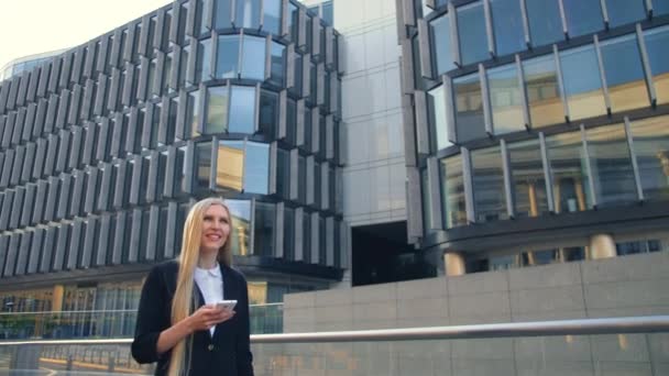 Smiling business woman with phone on street. Modern blond woman in elegant outfit holding phone and standing on street with modern architectural buildings. — Stock Video