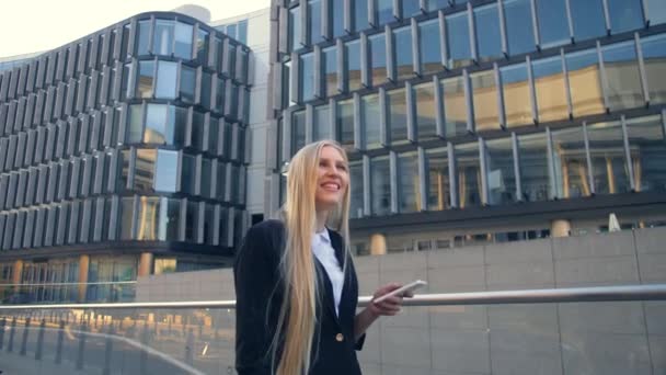 Glimlachende zakenvrouw met telefoon op straat. Moderne blonde vrouw in elegante outfit telefoon bedrijf en staande op straat met moderne architectonische gebouwen. — Stockvideo