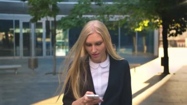 Açık havada telefon ile neşeli bir iş kadını. Akıllı telefon tutarak ve uzak gülümseme ile sokak arka plan üzerinde seyir siyah ceketli sarışın kadın içerik