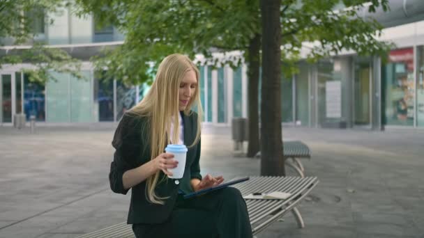 Ung affärskvinna med surfplatta och dricka kaffe. Moderna executive kvinna i kostym sitter på bänken utomhus surfing tablett och ha kopp kaffe. — Stockvideo