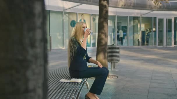 Mulher com café e comprimido no banco. Vista lateral da mulher formal sentada com tablet no banco e bebendo café olhando pensivamente longe no lazer . — Vídeo de Stock