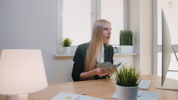 Kadın ofiste başarıyı kutluyor. Zarif sarışın kadın iş yerinde elinde bir tomar parayla oturmuş monitöre hayret ediyor, havaya para saçıyor ve mutlu bir şekilde evet hareketi yapıyor.. — Stok video