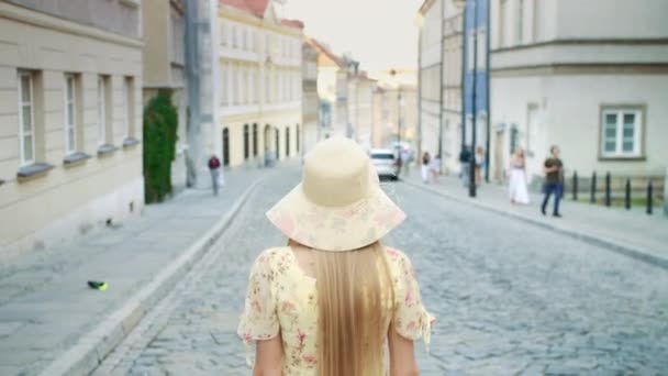 Mujer sonriente caminando por la calle. Mujer bonita alegre en sombrero blanco mirando hacia atrás a la cámara mientras camina por la ciudad . — Vídeo de stock