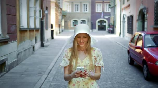 Kvinna textning på gatan. Gladlynt kvinna i vit hatt gå på gamla stan street och meddelandehantering med smartphone. — Stockvideo