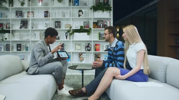 Hombre formal negro haciendo presentación con la tableta. Joven hombre afroamericano de traje sentado en la oficina en el sofá con un colega anónimo y haciendo la presentación con la tableta . — Vídeos de Stock