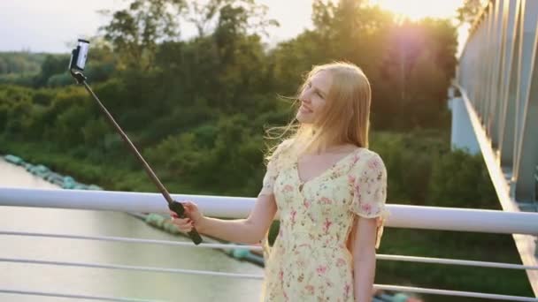 Woman taking selfie on bridge. Cheerful pretty blonde woman standing on pedestrian bridge and taking selfie with monopod in sunset. — Stock Video