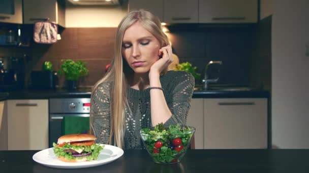 Νεαρή γυναίκα επιλέγοντας μεταξύ χάμπουργκερ και σαλάτα. Όμορφη νεαρή κοπέλα που βλέπουν νόστιμα burger και υγιεινή σαλάτα λαχανικών, ενώ κάθεται στο τραπέζι στην κομψή κουζίνα. — Αρχείο Βίντεο