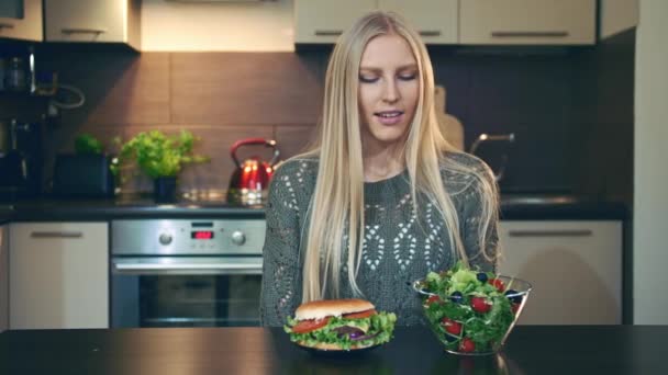 Девушка предпочитает гамбургер салату. Привлекательная молодая женщина выбирает здоровый гамбургер на завтрак, сидя за столом на стильной кухне . — стоковое видео