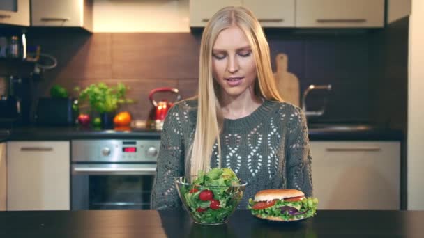 Young lady preferring hamburger to salad. Attractive young woman choosing to eat healthy hamburger for lunch while sitting at table in stylish kitchen and doing thumbs up gesture and looking at camera — Stock Video