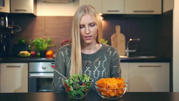 Young lady preferring salad to crisp. Beautiful young woman choosing to eat healthy crisp for supper while sitting at table in stylish kitchen and she eats these salad. — Stock Video