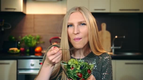 Glad kvinna som äter hälsosam sallad. Vacker ung kvinna njuter av hälsosam grönsakssallad och tittar på kameran medan du sitter i elegant kök. — Stockvideo