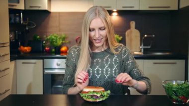 Genç kadın yemek fast food, hamburger şık mutfak masasında otururken. Genç Bayan hamburger salata için tercih.