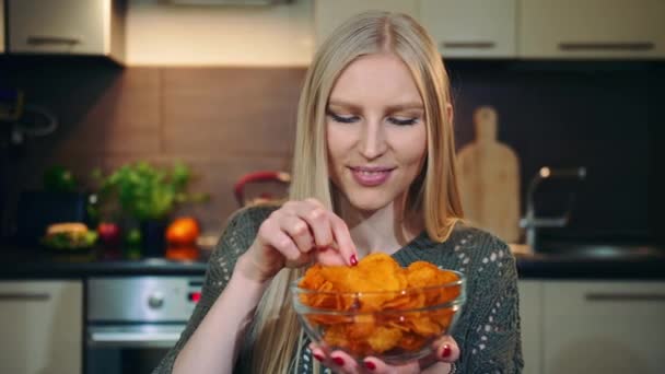 Glad kvinna som äter chips. Vacker ung kvinna njuter av potatischips och tittar på kameran medan du sitter i elegant kök. — Stockvideo