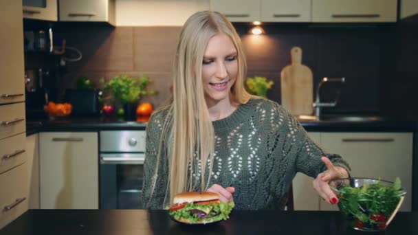 Young lady preferring hamburger to salad. Attractive young woman choosing to eat healthy hamburger for breakfast while sitting at table in stylish kitchen. — Stock Video