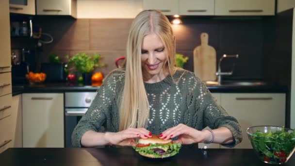 Jovem mulher comendo fast food, hambúrguer enquanto se senta à mesa na cozinha elegante . — Vídeo de Stock