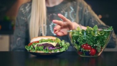 Hamburgeri salataya tercih eden genç bir bayan. Çekici genç bir kadın şık bir mutfakta masada otururken kahvaltıda sağlıklı hamburger yemeyi seçiyor..