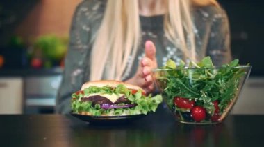 Hamburger için salata tercih kız. Çekici genç kadın şık mutfak masasında otururken akşam yemeği için sağlıklı salata yemek için tercih.