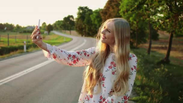 田舎道に近いビデオ通話の女性。若い女性の笑顔と現代のスマート フォンを使用してヨーロッパの田舎でヒッチハイクしながらビデオ通話にする. — ストック動画