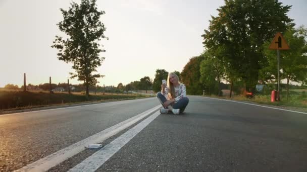 Mujer joven tomando selfie en la carretera. Encantadora jovencita sonriendo y posando para selfie mientras está sentada en la carretera en el campo de Europa . — Vídeos de Stock