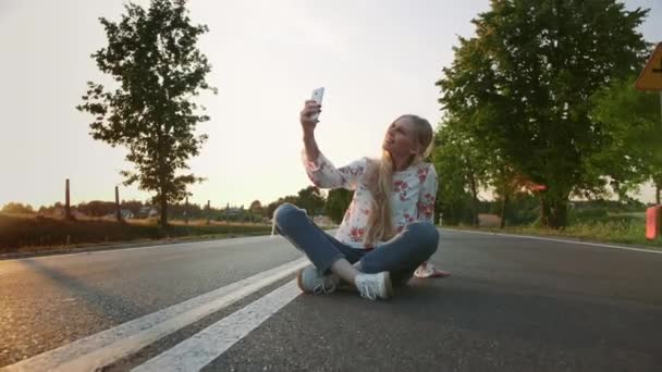 Ung kvinna tar selfie på väg. Härlig ung dam leende och poserar för selfie sittande på väg i Europa landsbygden. — Stockvideo