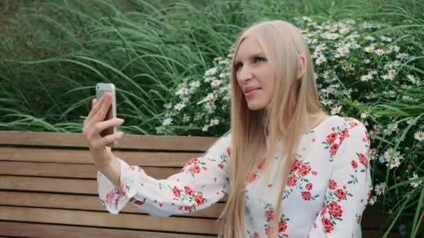 Woman making video call on green roof. Lovely young female using smartphone to make video call while sitting on bench on living roof of huge mall in USA. — Stock Video