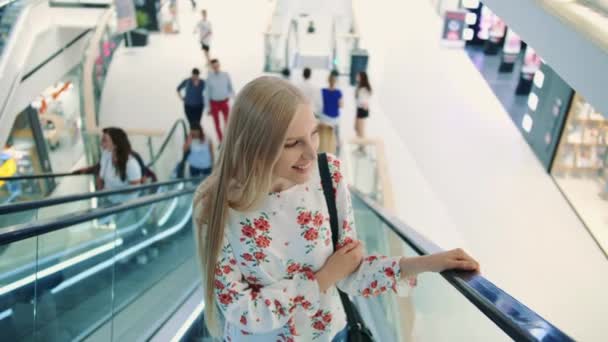 年轻的金发女人骑在自动扶梯上 购物中心里令人惊讶的女人。 在购物中心的一栋未来派建筑的背景下，身穿雅致服装的年轻女士乘坐自动扶梯. — 图库视频影像