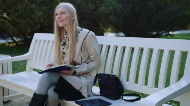 Krásná žena sedí na lavičce v podzimním parku, napíše do poznámkového bloku — Stock video