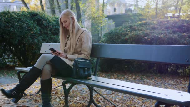 Menina bonita em jaqueta elegante usando smartphone no parque no dia ensolarado no parque de outono . — Vídeo de Stock