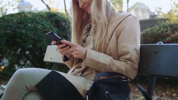 Bella ragazza in giacca elegante utilizzando smartphone nel parco nella giornata di sole nel parco autunnale . — Video Stock