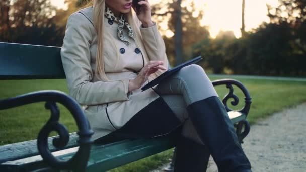 美丽的女孩在长凳上与平板电脑和手机坐在公园长椅上. — 图库视频影像