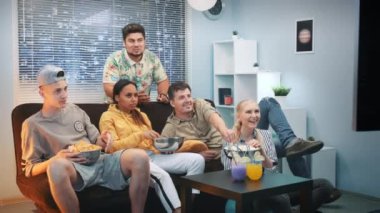 Heyecanlı arkadaşlar kanepede dinleniyor, TV izliyor ve evde atıştırmalık şeyler yiyorlar.