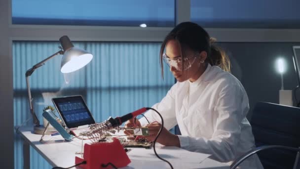 Primer plano medio del ingeniero de electrónica afroamericano en gafas protectoras comprobando placa base con probador de multímetro — Vídeo de stock