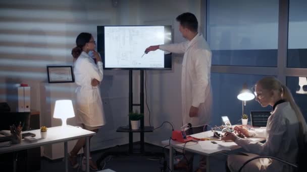 電子開発エンジニアのスマートチームは、研究室で大きなテレビ画面で何かを議論します — ストック動画
