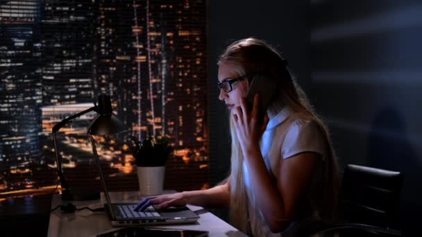 Молодая женщина-менеджер работает за компьютером и разговаривает с кем-то на смартфоне — стоковое видео