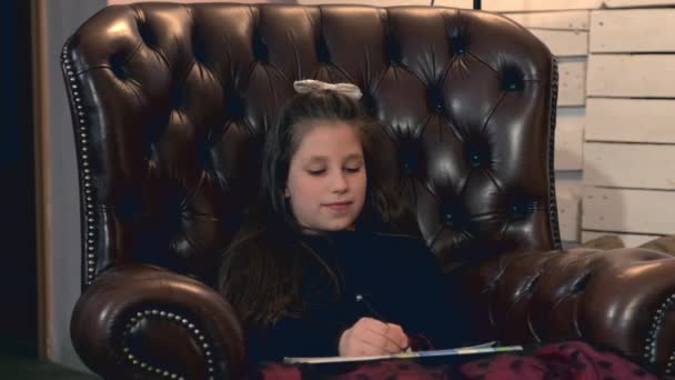 Küçük güzel kız bir deri koltukta oturan bir resim boyama — Stok video