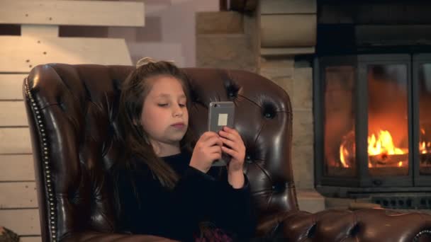 Liten söt flicka spelar spel på smartphone nära öppen spis — Stockvideo